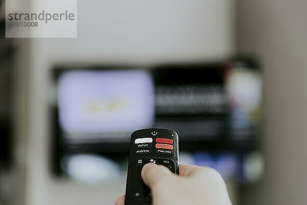 Hand holding remote control pointing to TV. Person watching smart television indoors. Blurred background. Colorful screen  copy space. Person browsing  looking for films online at home living room