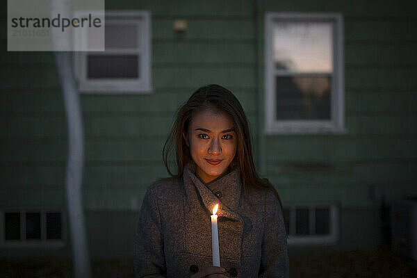 An Asian girl holds a candle in the dark in front of a small house.