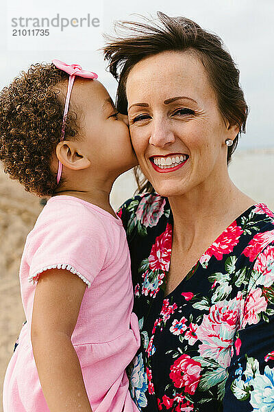 Toddler diverse girl kisses cheek of mother at beach