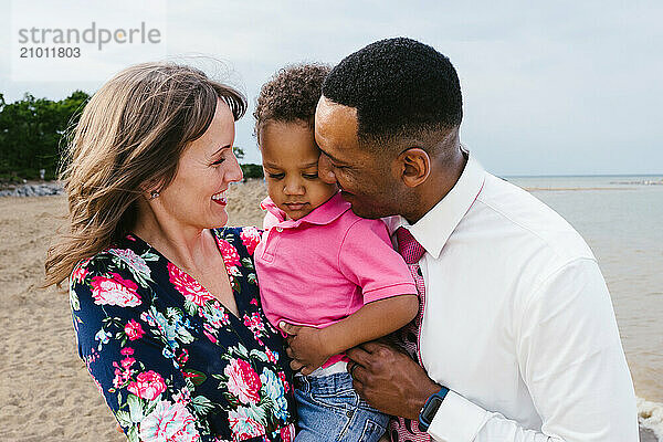 Dad and Mom hold toddler on the beach together as parents