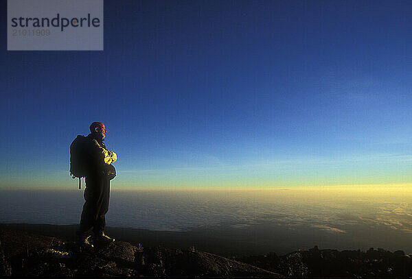 A man stands alone at sunrise as he looks down to the Tanzanian plains below Mount Kilimanjaro. John is attempting to climb to t