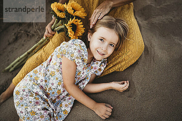 Girl lying on mom lap holding sunflowers at beach in summer