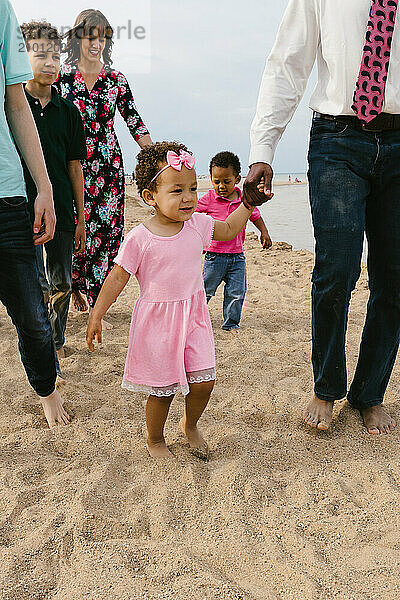 Toddler girl in pink dress walks with family along beach