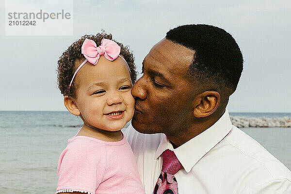 Black father kisses toddler daughter on cheek at the beach