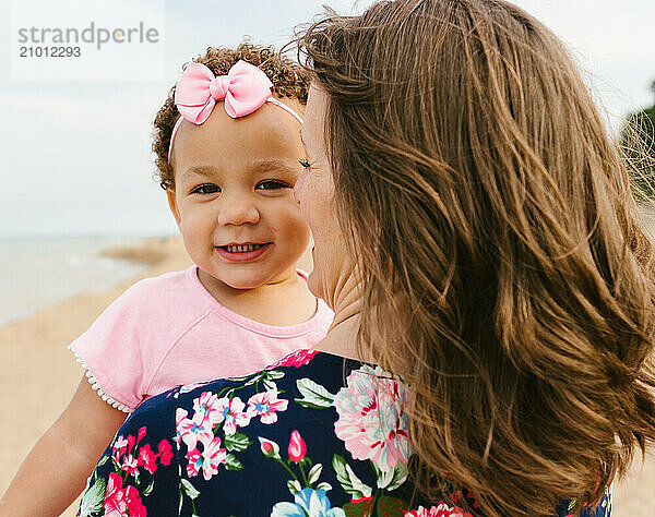 Toddler girl smiles over mothers shoulder on beach