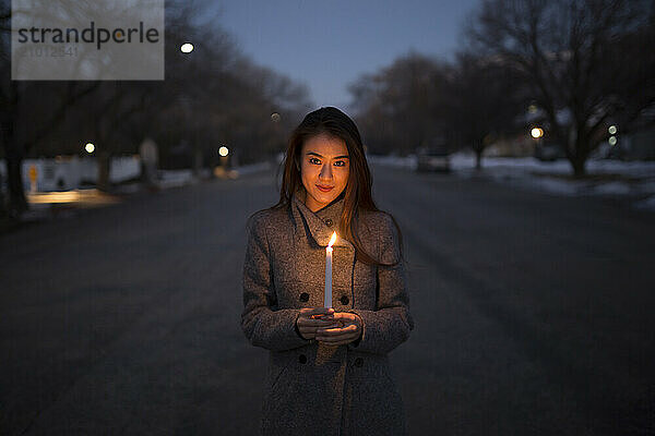 An Asian girl in a coat holds a candle at dusk in the road of a small town environment.