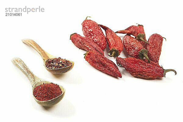 Red pepper powder and flakes on wooden spoons next to whole red peppers on a white background