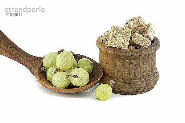 Gooseberries in a wooden spoon next to brown cane sugar cubes in wooden bowl  isolated on white background  berry jam  compote preparation