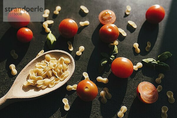 Red cherry tomatoes  green basil  butterfly vermicelli pasta  wooden spoon on gray background.