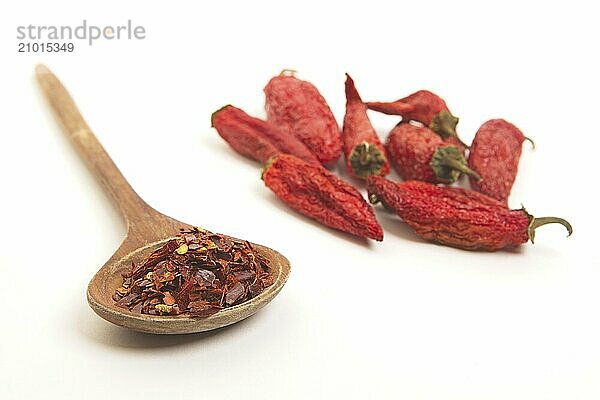 A still life photo of red pepper flakes on a wooden spoon next to shriveling red peppers on a white background