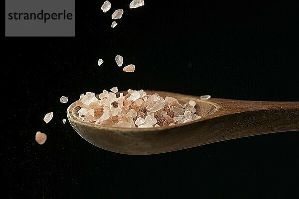 A close up photo a coarse pink himalayan sea salt falling down on the wooden spoon set against a black background