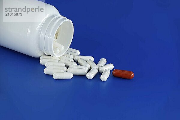 Overturned white medicine bottle with its contents white pills or capsules  spilled out onto a blue background. Among these white pills  a solitary red pill stands out