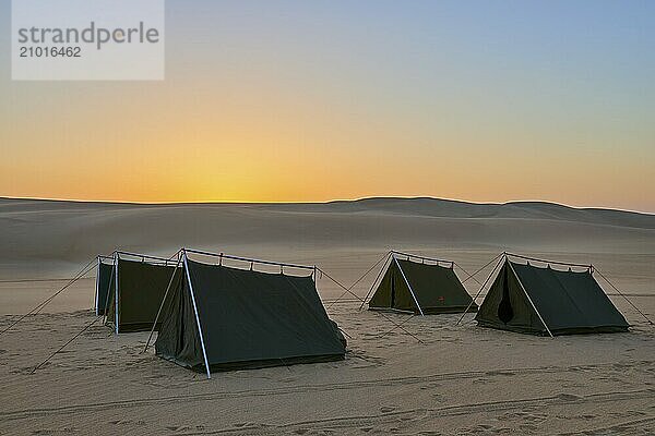 Four tents stand in a vast desert at dusk  surrounded by sand dunes under an orange-coloured sky  Matruh  Great Sand Sea  Libyan Desert  Sahara  Egypt  North Africa  Africa