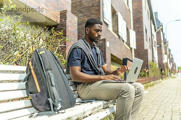 Side view portrait of a young african man working with laptop sitting on a city bench