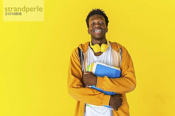 Studio portrait with yellow background of an african american cheerful student holding notebooks and folders with headphones on the neck