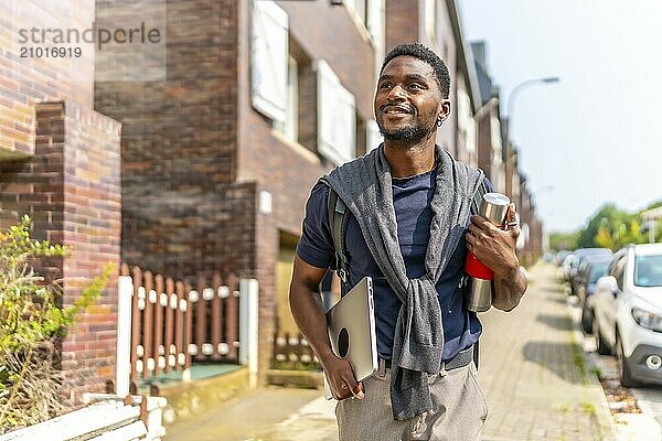 Happy african man carrying laptop and coffee thermos in the city on the way to work