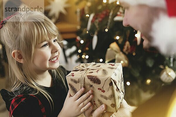 Young father in red Santa hat giving Christmas gift to his smiling daughter near decorated Christmas tree. Girl dressed in festive red-black Christmas outfit. They smiling  happy because of New Year