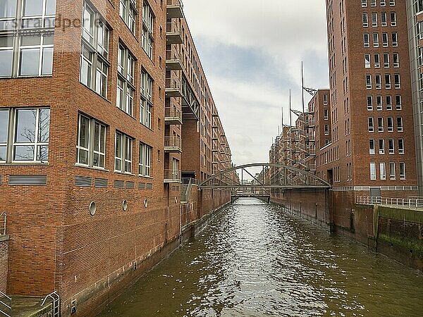 Narrow canal between brick buildings with a bridge under a cloudy sky  Hamburg  Germany  Europe