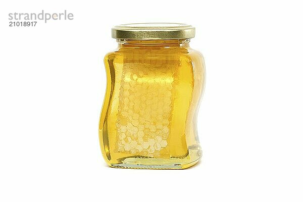 A glass jar with a golden lid contains honey with honeycomb filled almost to the top  against a white background