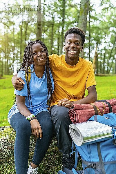 Vertical portrait of two young african friends sitting on fallen tree and resting during hiking