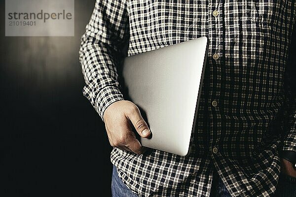 Man wearing casual clothes holds closed laptop  black blurred background.