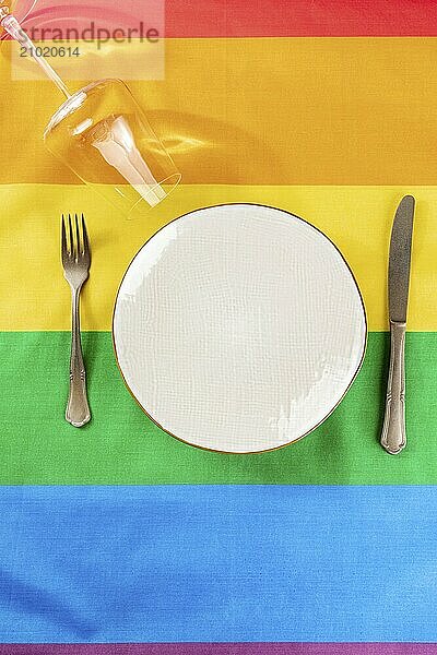 Gay dinner invitation design. A plate with cutlery and a wine glass  overhead flat lay shot on the LGBTQ rainbow flag texture