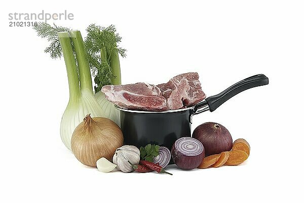 Black saucepan with pieces of raw pork atop. Around the saucepan are sliced vegetables which include onions  carrots  and garlic isolated on white background. Preparation of meat broth