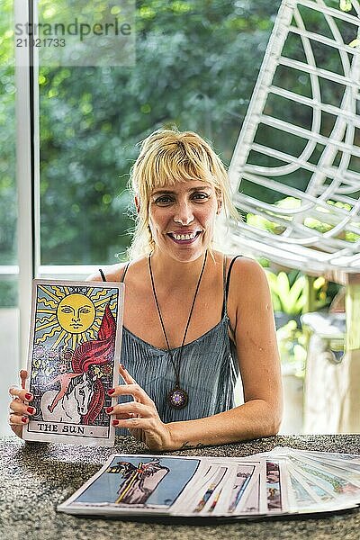 A tarot reader joyfully showing a large card of The Sun while looking at the camera