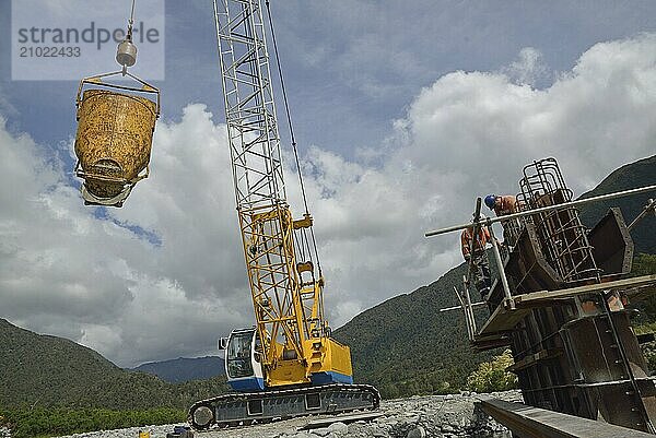 A crane driver swings across a bucket of cement for builders pouring a pylon for a concrete bridge over a small river in Westland  New Zealand  Oceania