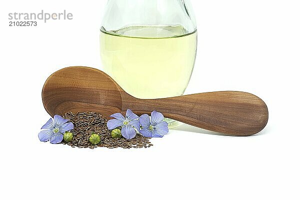 Wooden spoon filled with small brown flax seeds and a blue flax plant flowers alongside linseed oil in glass decanter isolated on white background