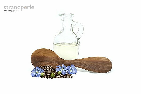 Wooden spoon filled with small brown flax seeds and a blue flax plant flowers alongside linseed oil in glass decanter isolated on white background