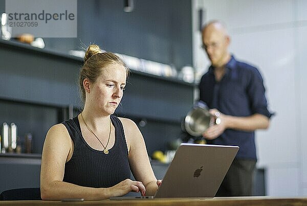 Symbolic photo on the subject of division of labour for couples in the household. A woman sits at a laptop in a kitchen while a man washes dishes in the background. Berlin  13.08.2024