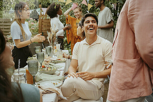 Smiling young man sitting at dining table and talking with friends in back yard party