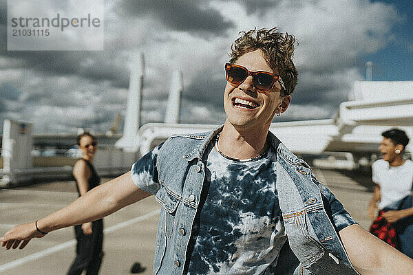 Portrait of cheerful young man in sunglasses with tattooed arm enjoying with friends in parking lot