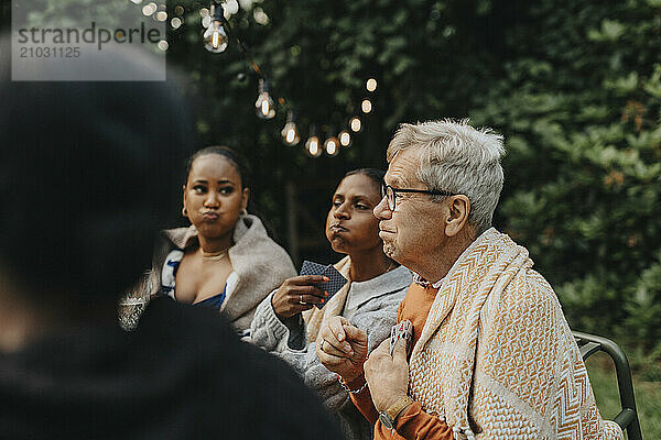 Senior man making face with family members and playing cards at garden party