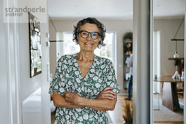 Portrait of smiling senior woman standing with arms crossed at home