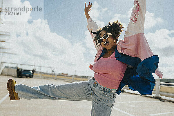 Cheerful curly hair woman in sunglasses and letterman jacket dancing at parking lot under cloudy sky