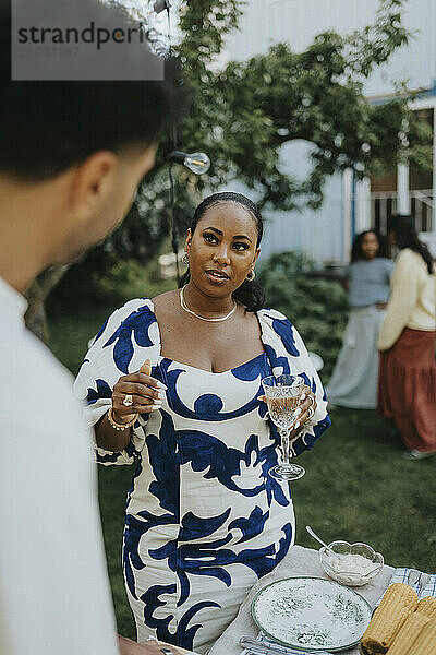 Mid adult woman holding glass while standing near table and discussing with male friend at social gathering