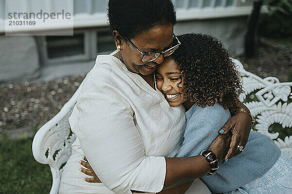 Smiling grandmother embracing granddaughter while sitting on bench at back yard