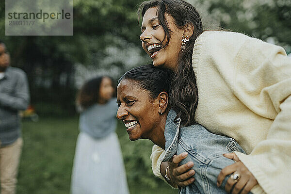 Cheerful mature woman piggybacking daughter at social gathering with family in back yard