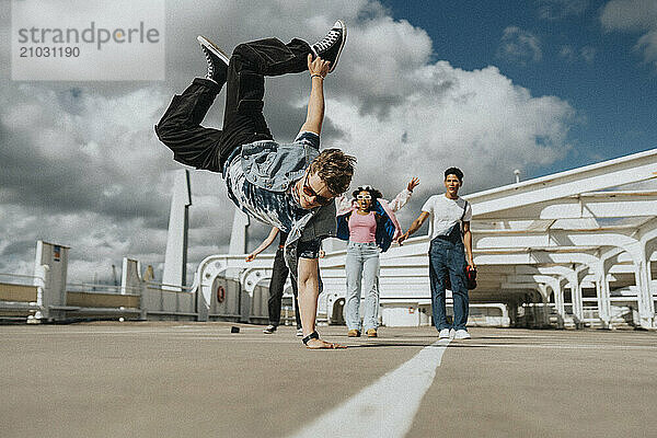 Athletic young man doing handstand while male and female friends cheering in parking lot under cloudy sky