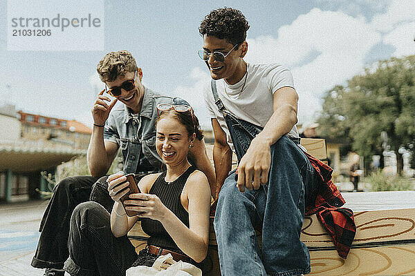 Happy young woman using smart phone with fashionable male friends while sitting at street