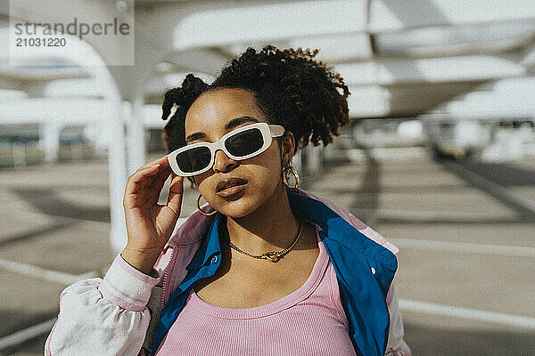 Portrait of fashionable curly hair woman holding sunglasses at parking lot in sunlight