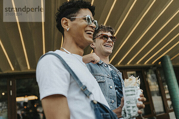 Low angle view of happy young man in sunglasses leaning on male friend holding wrap sandwich outside shop