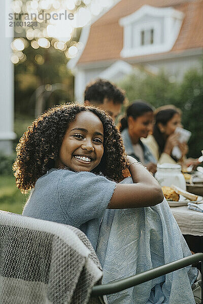 Portrait of happy curly hair girl sitting on chair at social gathering in back yard