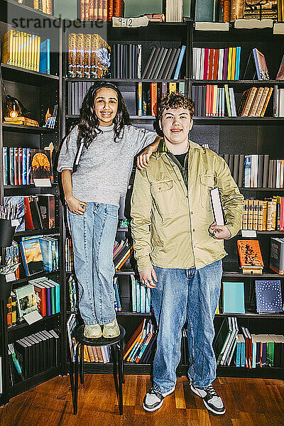 Smiling girl standing next to male friend holding book near shelf in bookstore