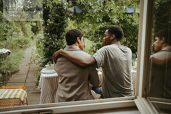 Rear view of young man sitting with arm around male friend near window