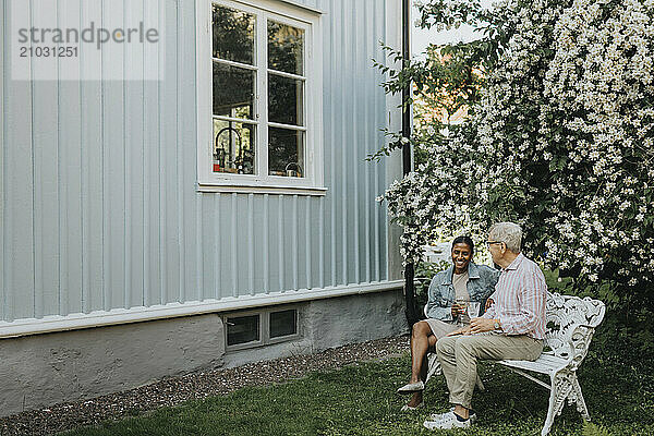 Happy mature woman discussing with senior family member and sitting on bench at back yard