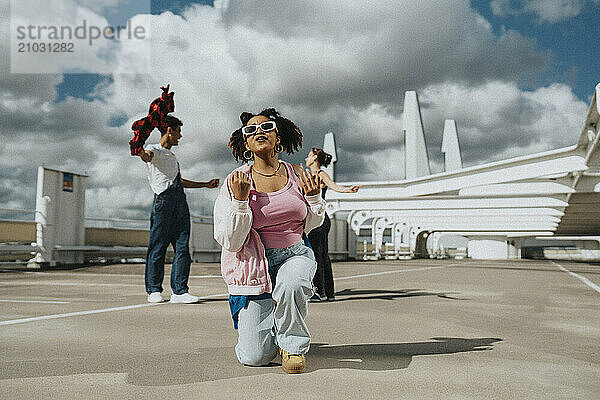 Carefree fashionable young woman kneeling and singing in parking lot under cloudy sky