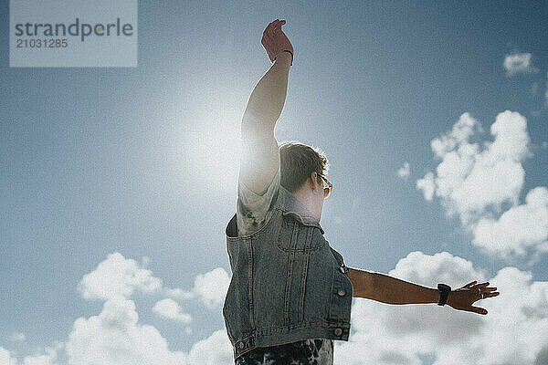 Low angle view of young man with tattooed arm and denim jacket dancing under cloudy sky at sunny day